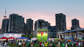 People in soccer jerseys at an outdoor festival gathered around a big screen watching a soccer game.