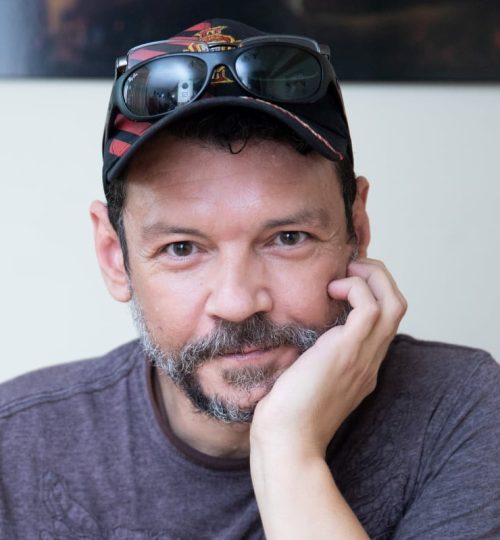 Headshot of man wearing black baseball cap with sunglasses on the cap.