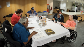 Residents seated around a table reading song sheets during a group music activity, with staff support and refreshments on the table.