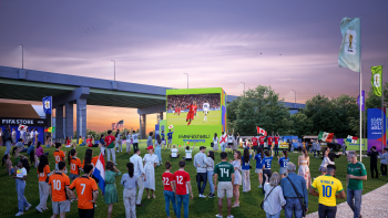 People in soccer jerseys at an outdoor festival gathered around a big screen watching a soccer game.