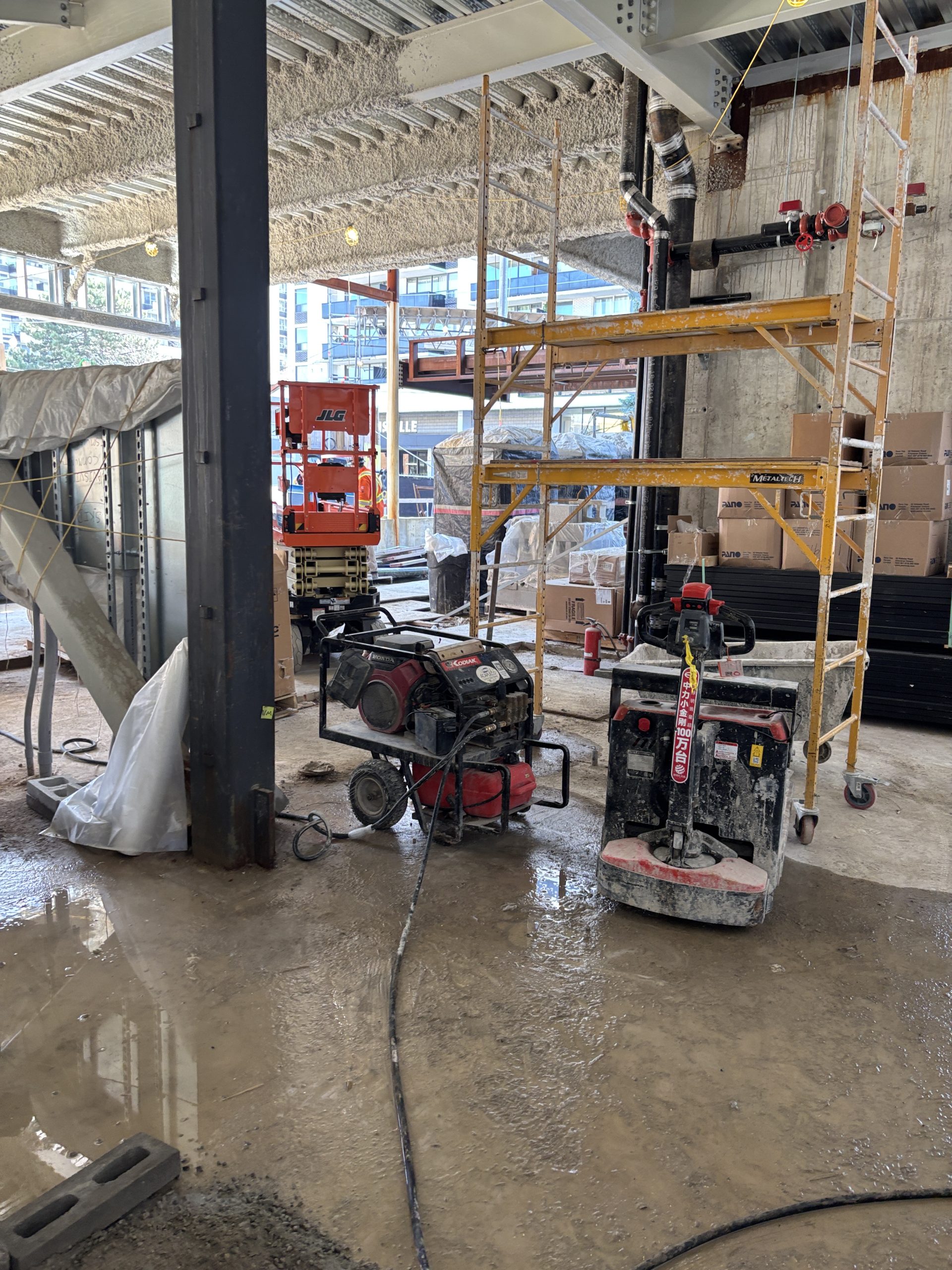 The interior of Davisville Community and Aquatic Centre under construction with concrete walls and exposed beams along the ceiling. Boxes and materials wrapped in plastic coverings surround equipment such as a scissor lift and a rolling scaffold.