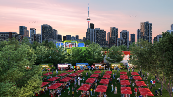 Red umbrellas cover picnic tables at an outdoor festival with a large viewing screen showing soccer in the background.