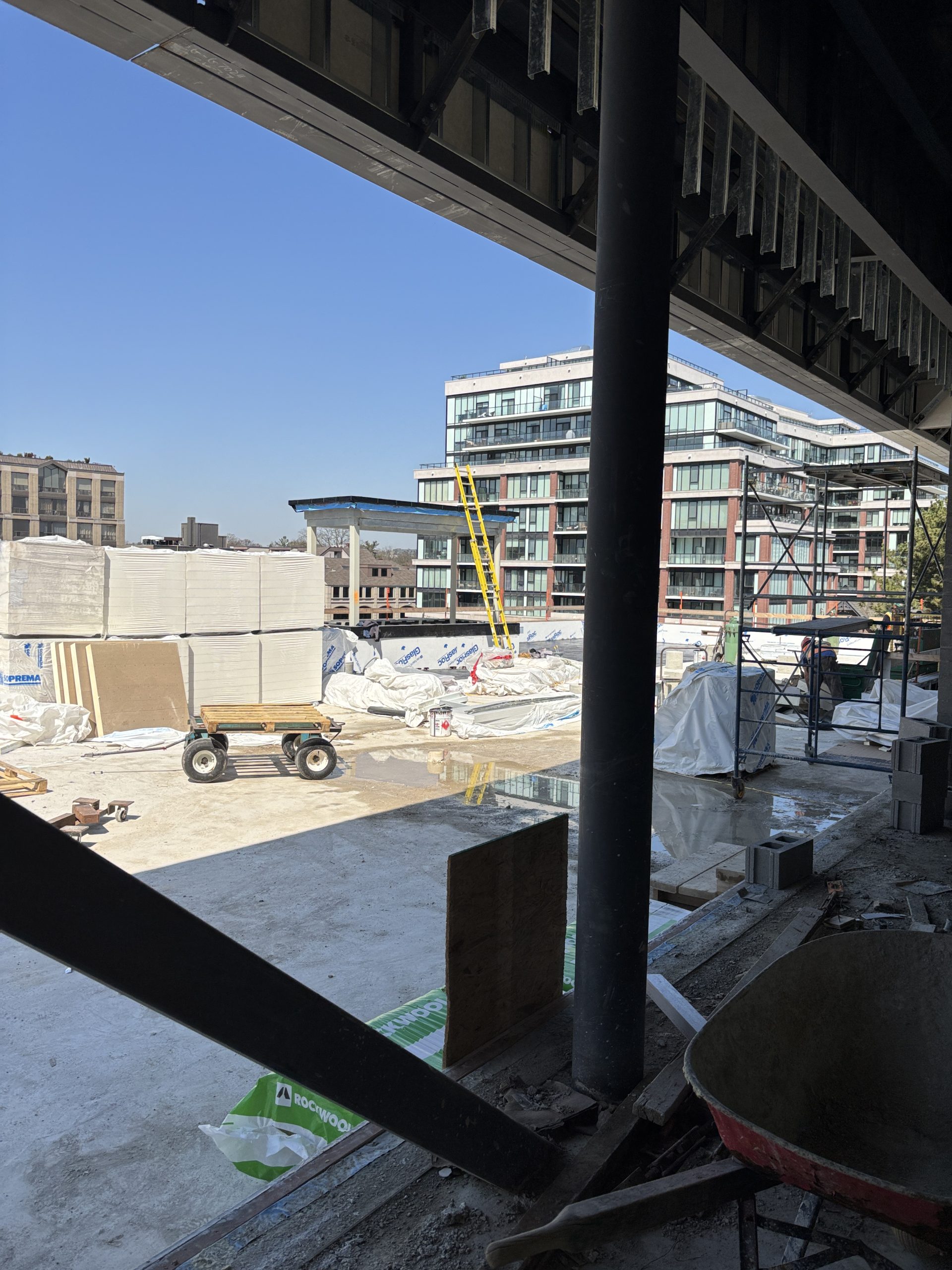 View from a partially enclosed construction area for Davisville Community and Aquatic Centre framed by steel beams. Wrapped building materials, scaffolding and equipment are scattered across a concrete surface outside. Residential buildings are visible in the background under a clear blue sky.