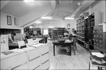 Image depicts people in archival reading room, with desks, bookshelves and filing cabinets.