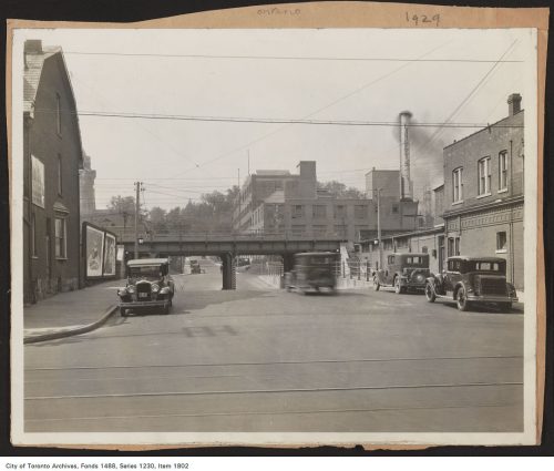 Image depicts monochrome street view with cars, stores and factory.
