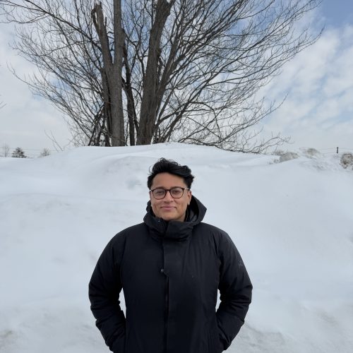 Photograph of man wearing black winter puffer jacket in front of snowed hill
