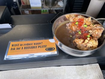 A reusable bowl full of food with a sign beside it reading want to reduce waste? Ask for a reusable plate!