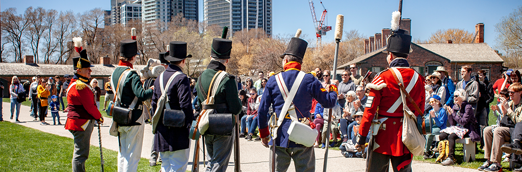 Historical soldiers wearing different uniforms stand in a line facing an onlooking crowd