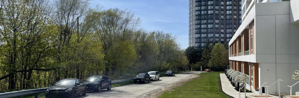 An eye-level view looking west along the extension of Hickory Tree Road at 10 Wilby Crescent. On the right side is a recently constructed multi-storey residential building with townhouse entrances, balconies, and a narrow lawn frontage. The image documents the future park access and surrounding site context.