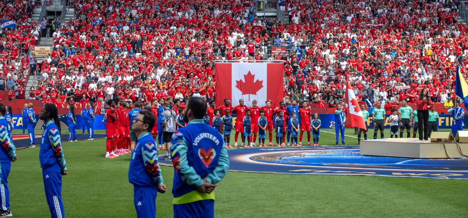 Opening ceremony at stadium. Singer stands on platform on right. Soccer team stands with children in front of Canadian flag. Official volunteers stand in arc. Audience in tiered seating in background.