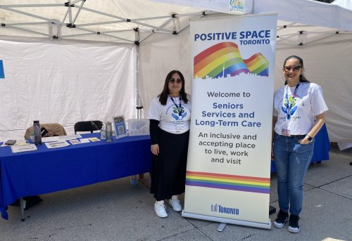 Two staff stand at an outdoor booth beside a banner reading “Positive Space Toronto – Seniors Services and Long-Term Care.”