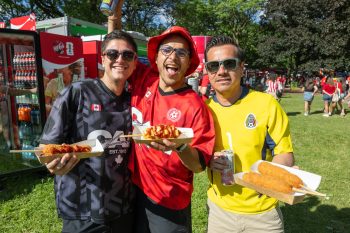 Three people wearing different soccer jerseys smile to camera, holding corn dogs