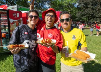 Three people wearing different soccer jerseys smile to camera, holding corn dogs