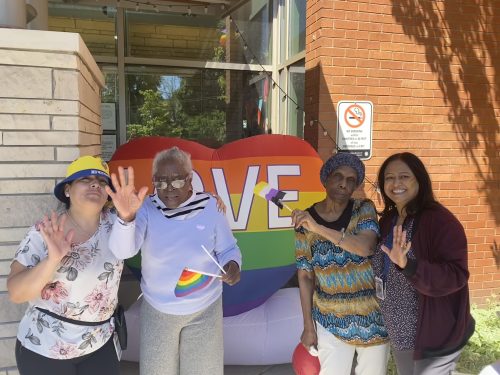 Four people standing outdoors in front of a rainbow heart display with the word “LOVE,” waving and holding small rainbow items.
