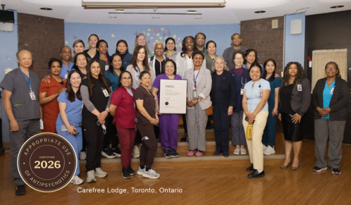 Large group of staff standing together indoors, holding a framed recognition certificate at Carefree Lodge in Toronto. On the bottom left of the image there is a medal, that says "Appropriate Use of Antipsychotics Certified 2026."