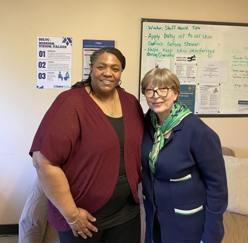 Two people stand side by side in an indoor office or staff area, posing for a photo. Behind them is a wall-mounted whiteboard with handwritten notes titled “Winter: Staff Health Tips,” along with printed notices and contact information. A poster to the left reads “SSLTC – Mission, Vision, Values.”