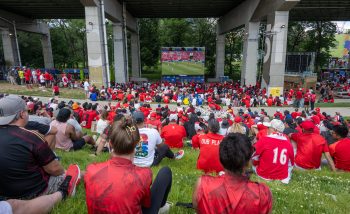 Soccer fans seated on hill watch match on screen underneath Gardiner Expressway.
