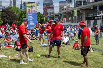 Three people wearing jersey kick soccer ball to each other outside on grass surrounded by festival goers.