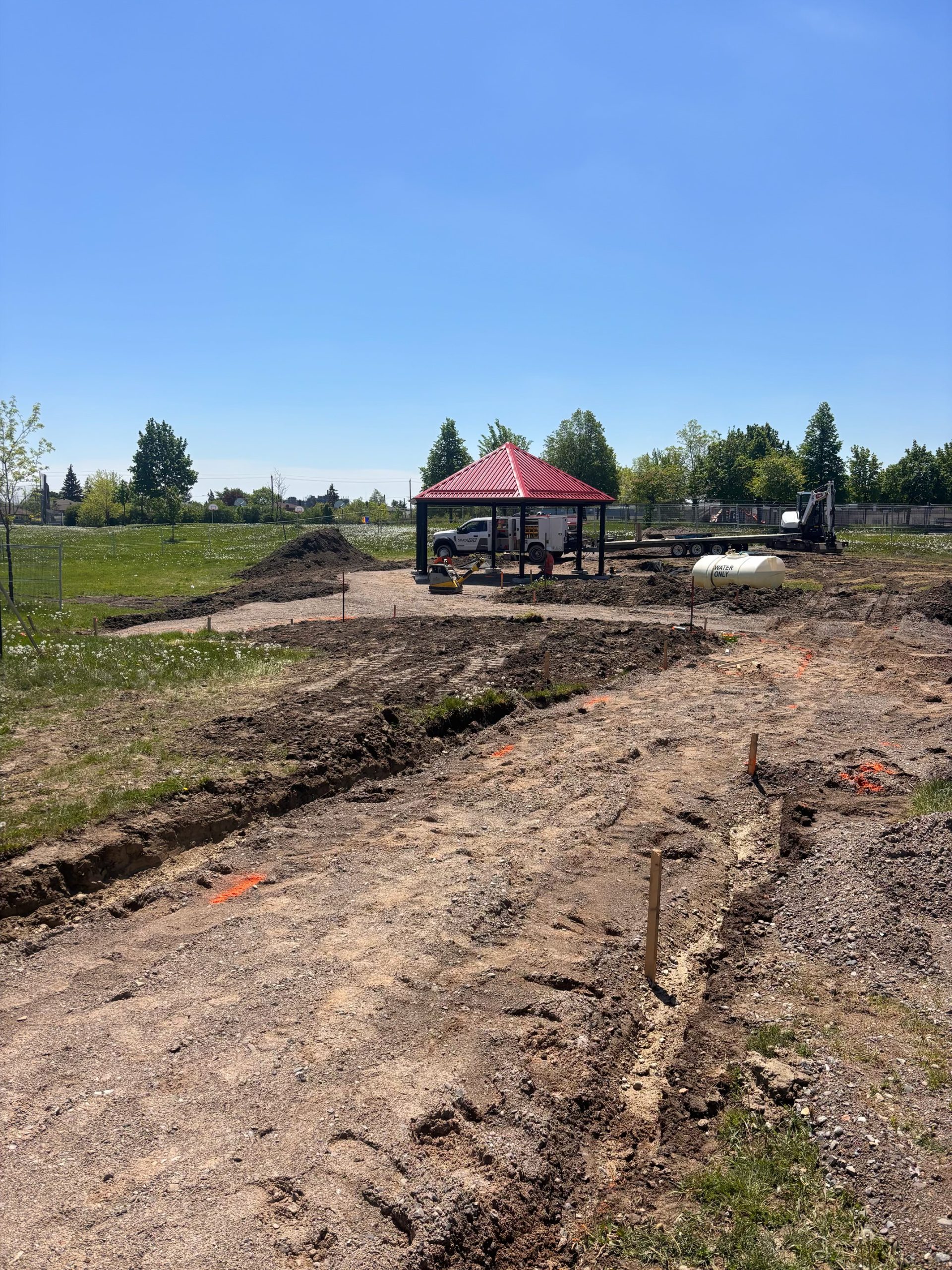 Construction site with a partially built shade shelter in the centre, surrounded by excavated dirt. Construction equipment, including a truck, trailer and excavator, is parked nearby. The site is bordered by an open grassy field and trees in the distance. 