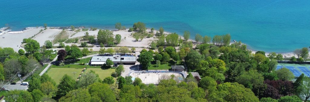 Aerial view of a Kew Gardens along Lake Ontario. Centered in the frame is an artificial ice rink being used as an outdoor recreation space during the summer months, surrounded by mature trees, open green space, and park pathways, with a sandy beach and shoreline visible in the background.