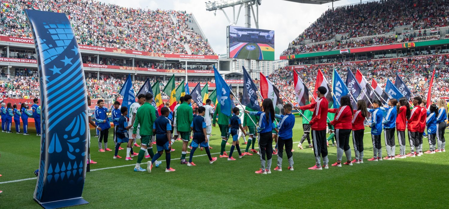 Soccer players walk between two lines of flag bearers in a stadium with tiers of seating filled with fans