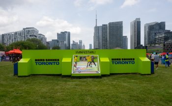 Booth with people playing soccer on lawn in front of city skyline. French text on booth nous sommes Toronto