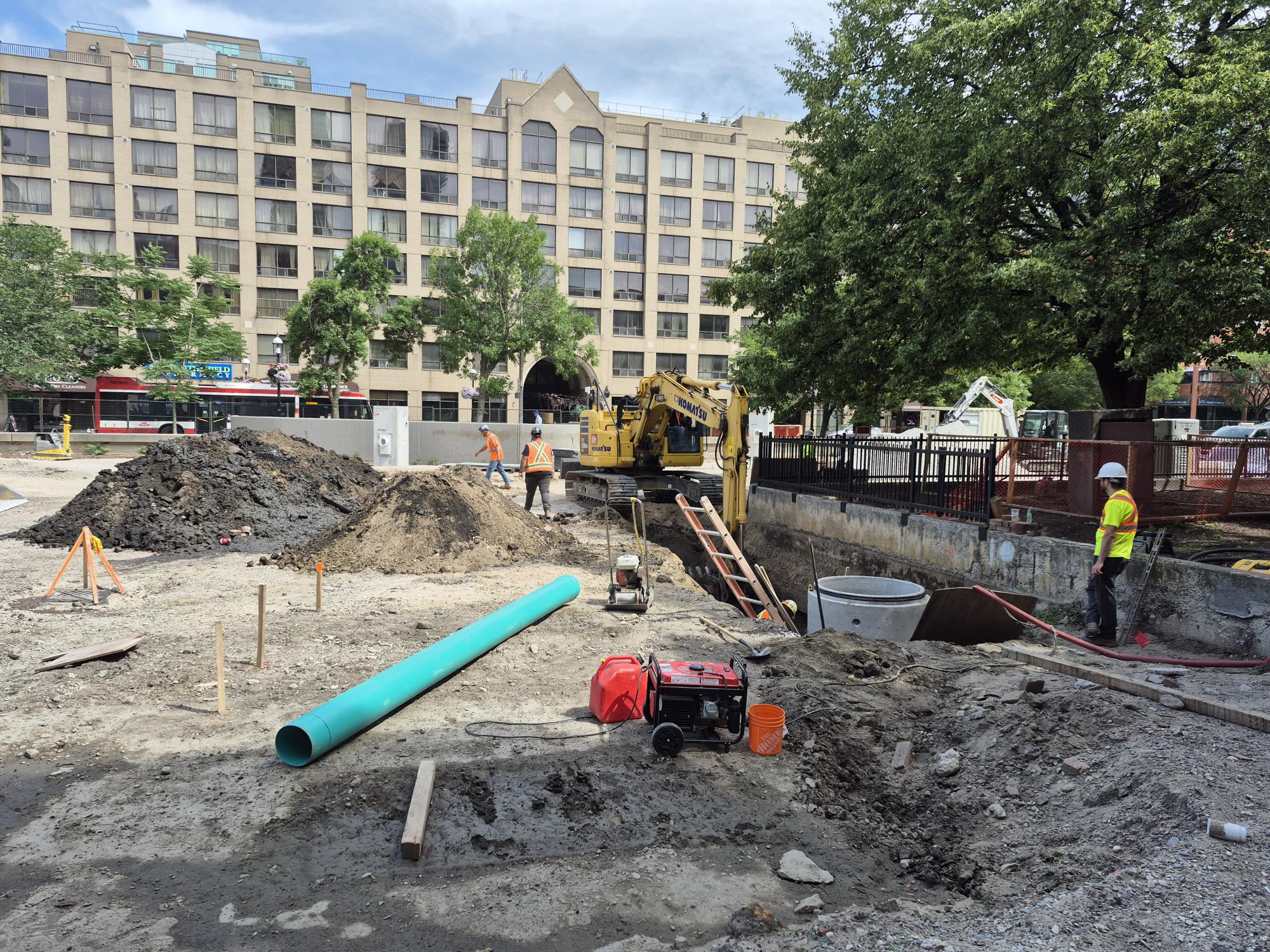 Construction activity in an urban park setting with excavation work underway. Workers in safety gear stand near a trench. Scattered around them are piles of soil and equipment such as a ladder, pipe, generator and compactor. Mature trees surround the site, and a residential building is visible in the background.