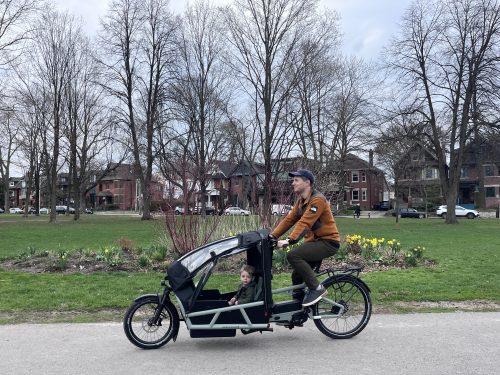 An adult rides a black cargo bicycle with a covered front passenger compartment carrying a young child along a paved path through a neighborhood park. 