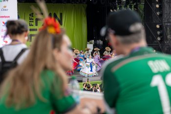 Dancers on stage as seen through the space between the shoulders two audience members wearing green soccer jerseys