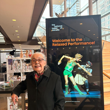 Older adult standing beside a digital display at the National Ballet of Canada reading “Welcome to the Relaxed Performance.”