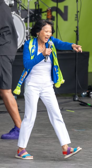 Mayor Olivia Chow wears a FIFA Toronto 2026 scarf and stands on stage, speaking into microphone and points to the right
