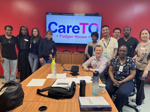 Care team members stand and sit around a meeting table during onboarding, with a screen displaying “CareTO Fudger House” behind them.