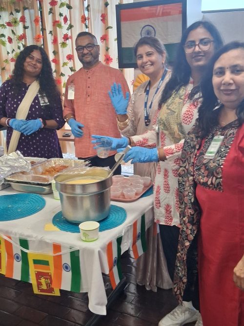 Staff wearing gloves serve food and stand behind a decorated table with dishes and small cups, in a room decorated with Indian flags and flowers.