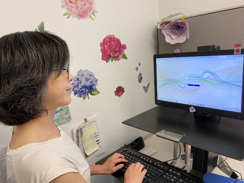 Staff member typing on a computer at a workstation, with a login screen visible on the monitor and floral wall decorations nearby.
