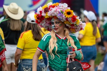 Fan wears floral wide-brimmed hat and Mexico jersey and has face painted with flag and skull
