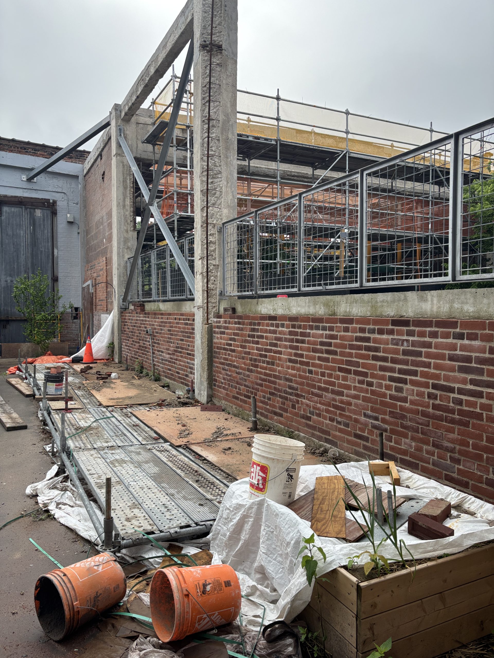 A construction site at Wychwood Barns Park beside a brick building with concrete columns and metal framing supporting a partially open structure. Scaffolding surrounds the building’s upper level, while the ground is cluttered with construction materials.