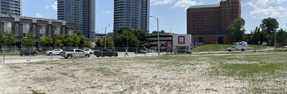 A large open area with sand and patches of grass with a tall metal fence around the perimeter. Two condos and residential homes can be seen in the background, adjacent to the site