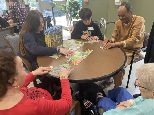 Residents and students gather around a table in a common area, working together on a large jigsaw puzzle with colourful bird images.