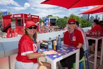 Two people seated at patio smile to camera. Festival crowd and booth in background.