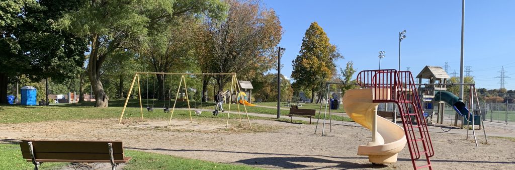 The existing playground at McNicoll Park. In the foreground, a wooden bench sits alongside a playground featuring a swing and slide. Across the sidewalk, a senior and junior play tower is visible on the opposite side.