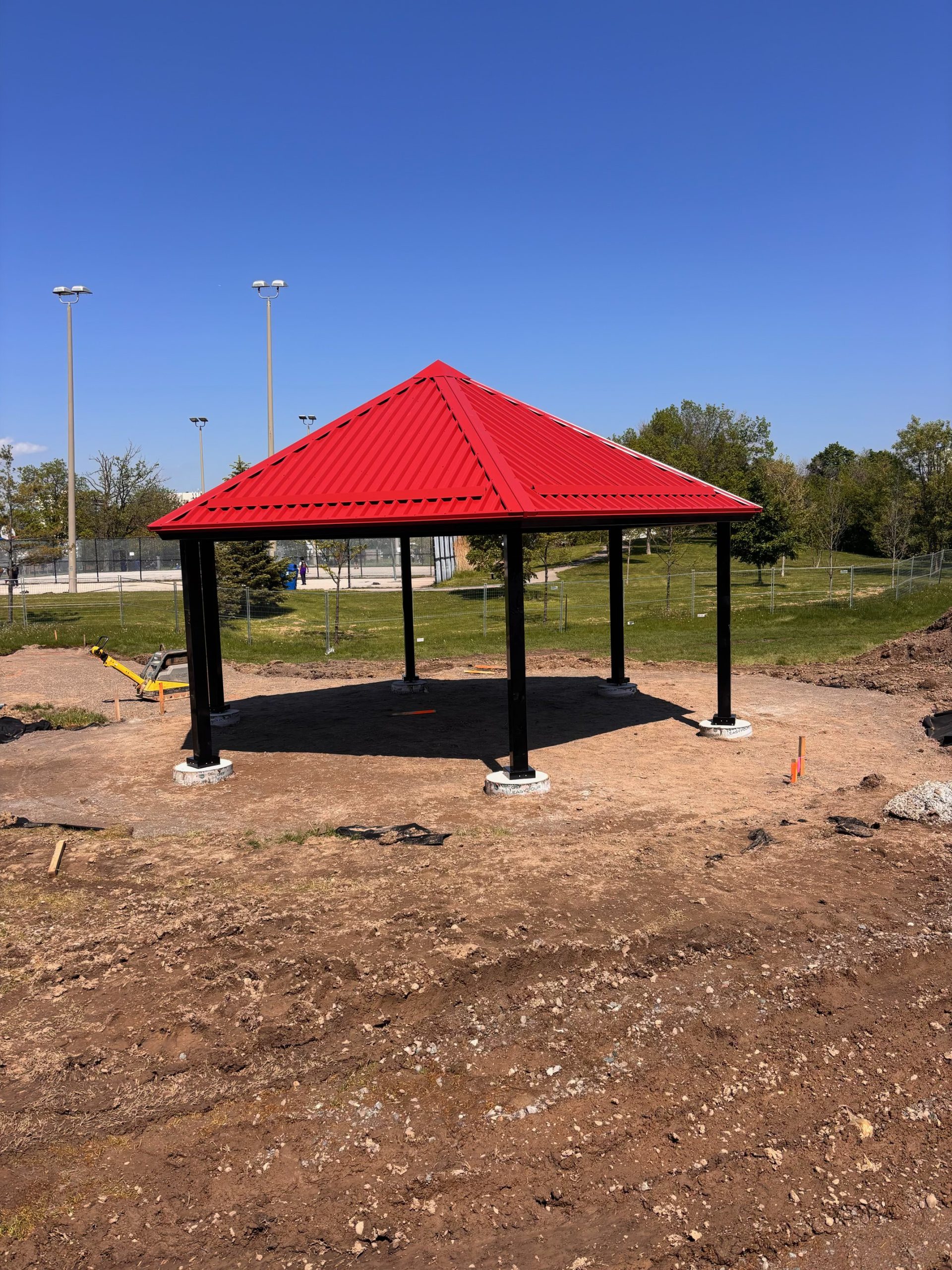 A shade shelter with a red roof and black posts under construction in a park, surrounded by dirt and graded ground. Safety fencing encloses the site. Trees and sports courts are visible in the background.