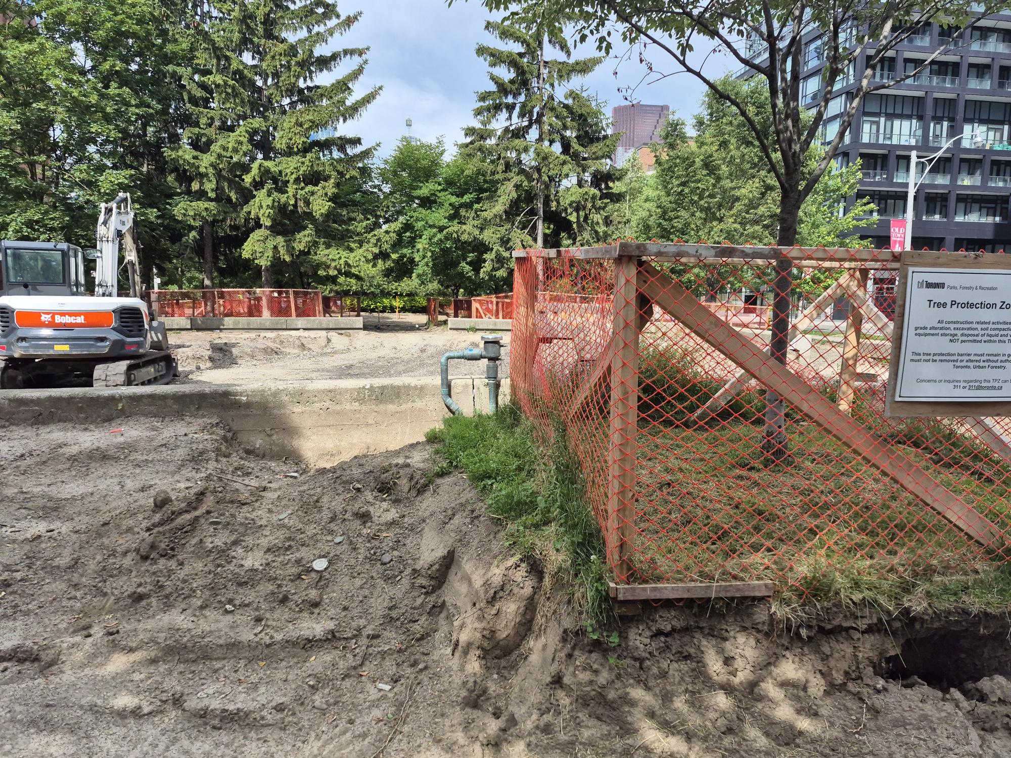 Construction site in a park area featuring an excavator and piles of exposed soil. The site is enclosed by orange safety fencing and surrounded by mature trees. Tree protection fencing is visible in the foreground.