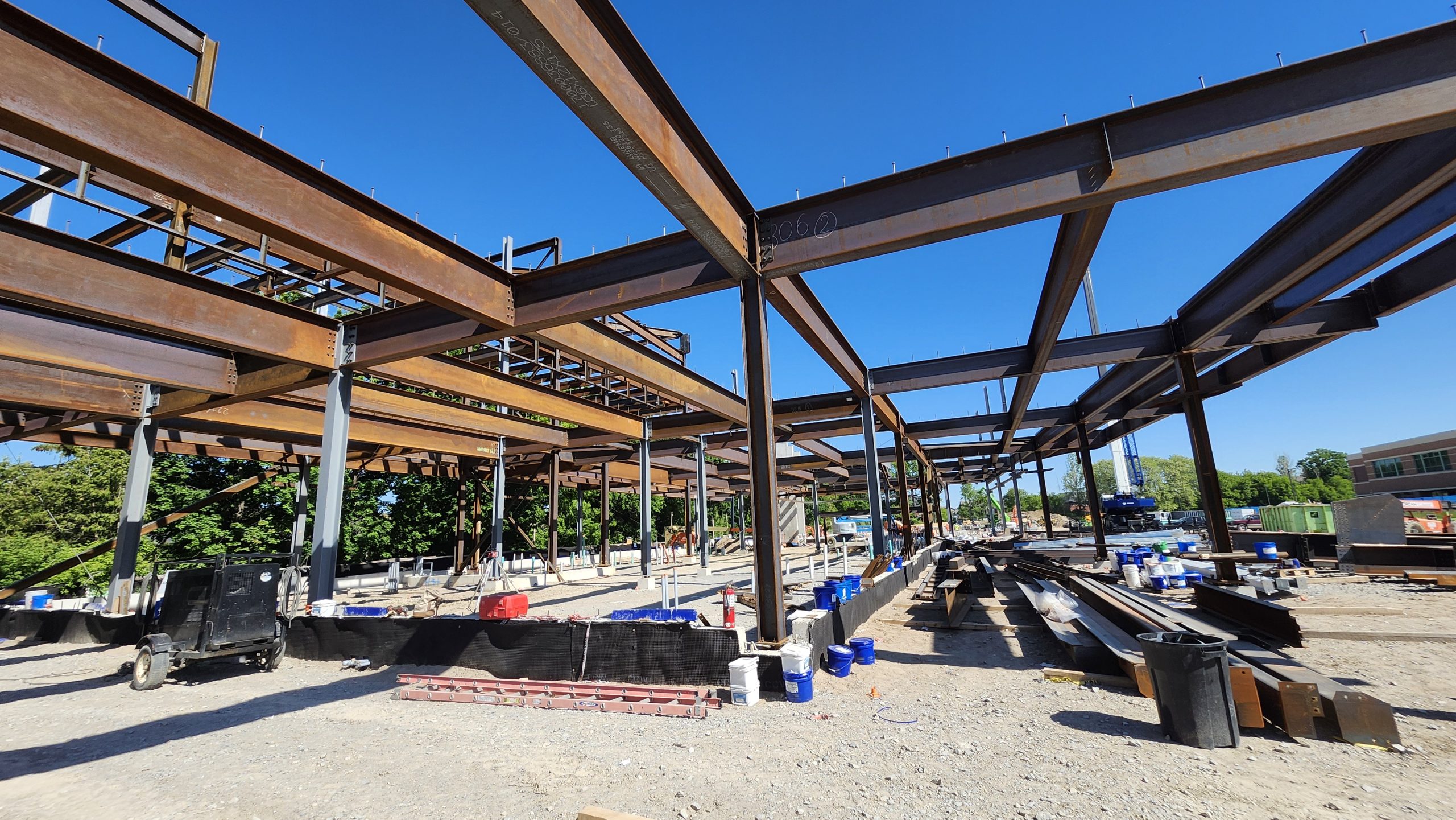 View of the structural steel erection for the new community centre from the north, facing the future main entrance and underneath the future gymnasium. The ground is covered with grave and scattered construction materials. A tall crane is visible in the background.