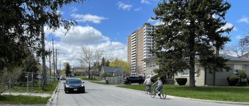 A photo of two people on bikes and a few cars, on a street in West Agincourt.