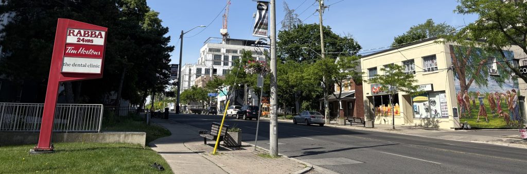 Photo with view facing west on Dundas Street West showing buildings with storefronts, street trees, and signage on the north and south sides of the street, a mural on the side main wall of one of the buildings on the north side of the street, and a mid-rise building under construction.