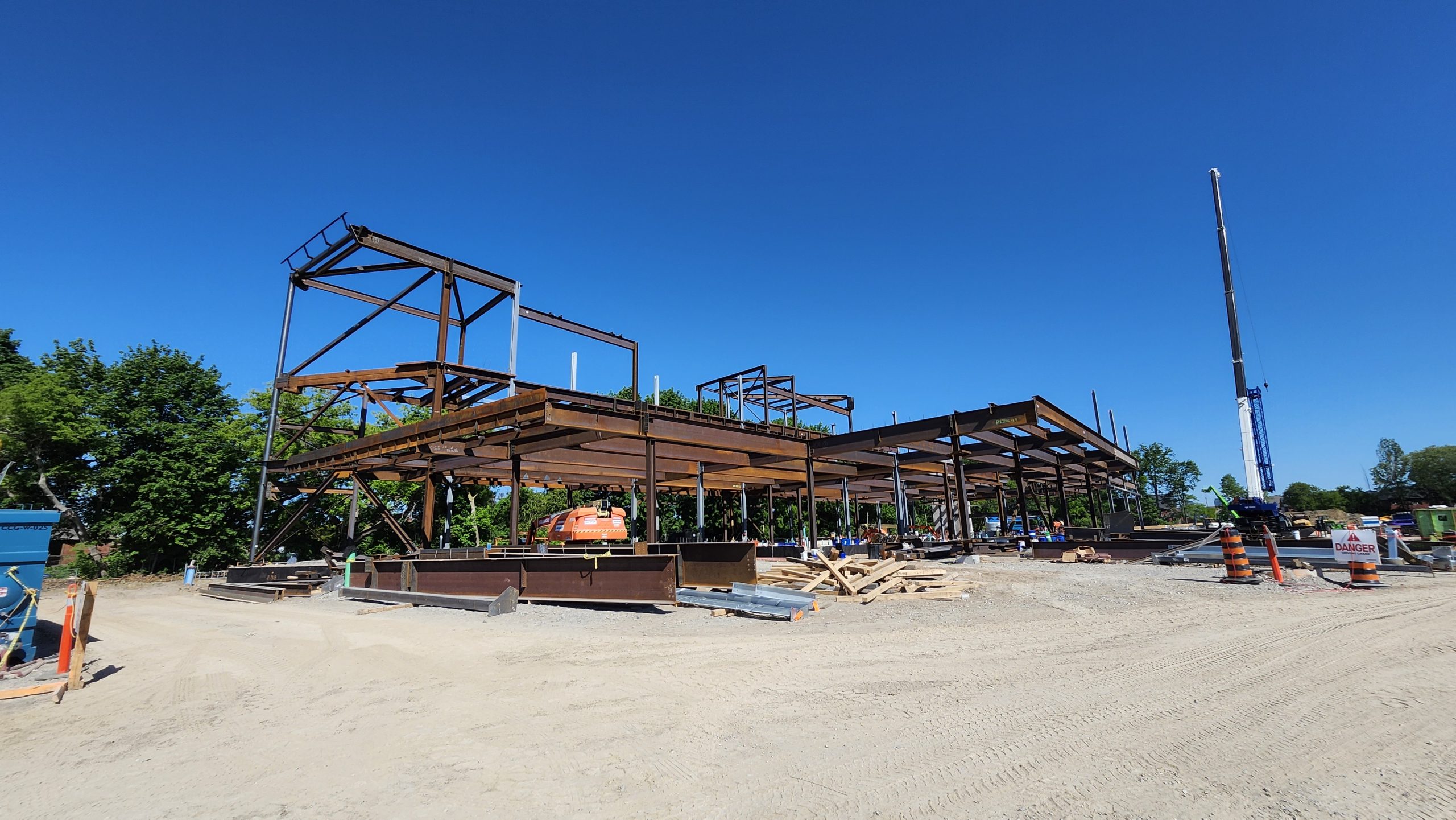 View of the overall structural steel erection for the new community centre from the northwest. Stacked materials and equipment surround the large steel structure. A tall crane stands to the right near orange safety cones.