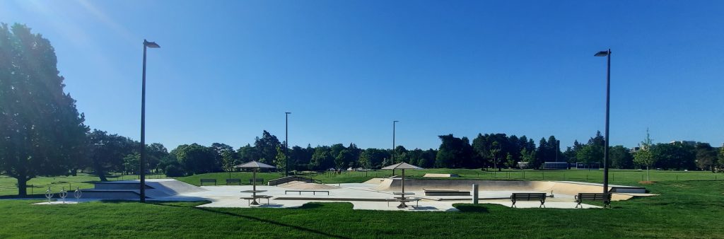 Wide view of an outdoor skatepark in a large grassy park featuring concrete ramps, rails and platforms. Benches, shade structures and tall lampposts are placed along the edge of the skate area.