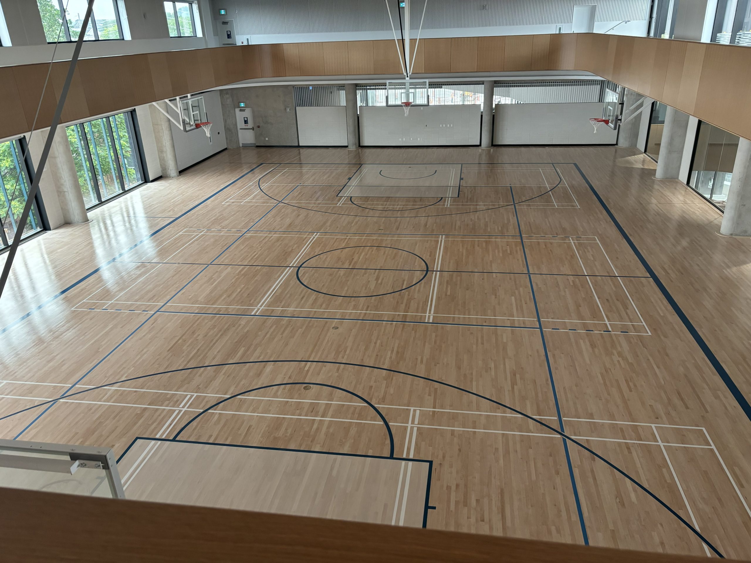 View of the gym in the community recreation centre from the upper-level running track. Court lines are painted on the wood floor, and basketball hoops are installed.
