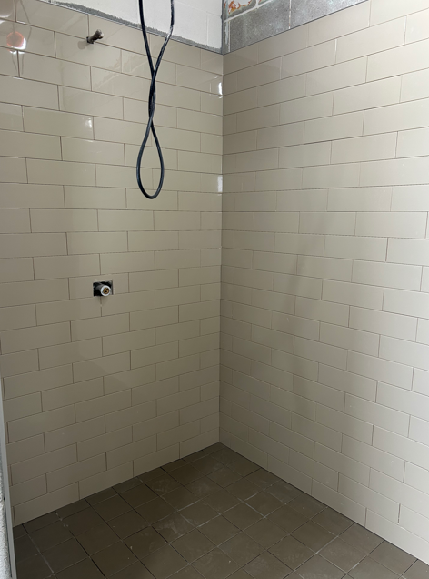 Completed shower stall with light beige subway tile walls and dark grey tile flooring, with an electrical outlet and hanging wire visible.