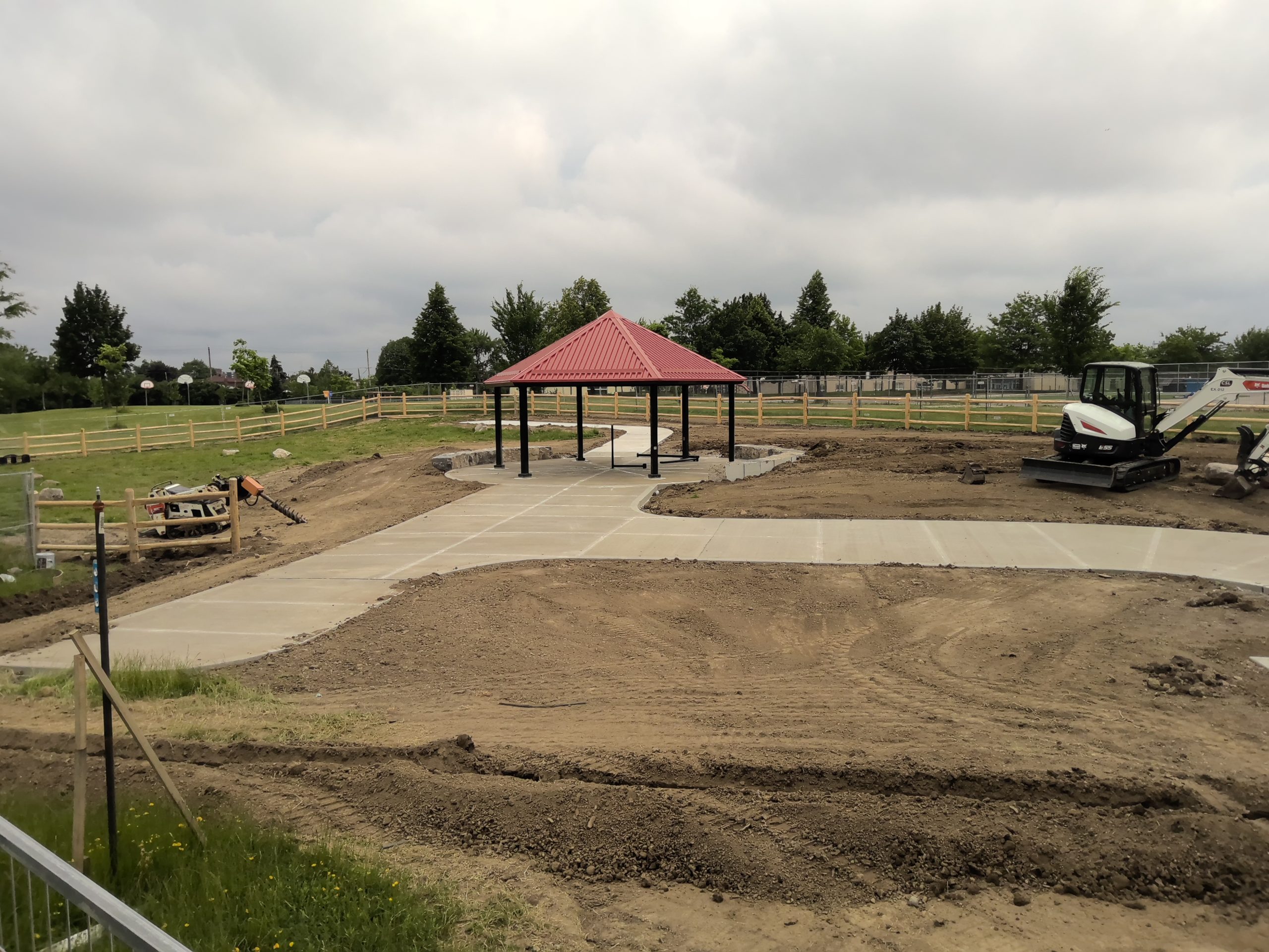 Construction site for an off-leash area with freshly paved concrete pathways that lead to a shade structure with a red roof. Landscaping is ongoing in areas with exposed soil. A wooden fence encloses the site, with trees, open fields and outdoor basketball courts visible in the background.